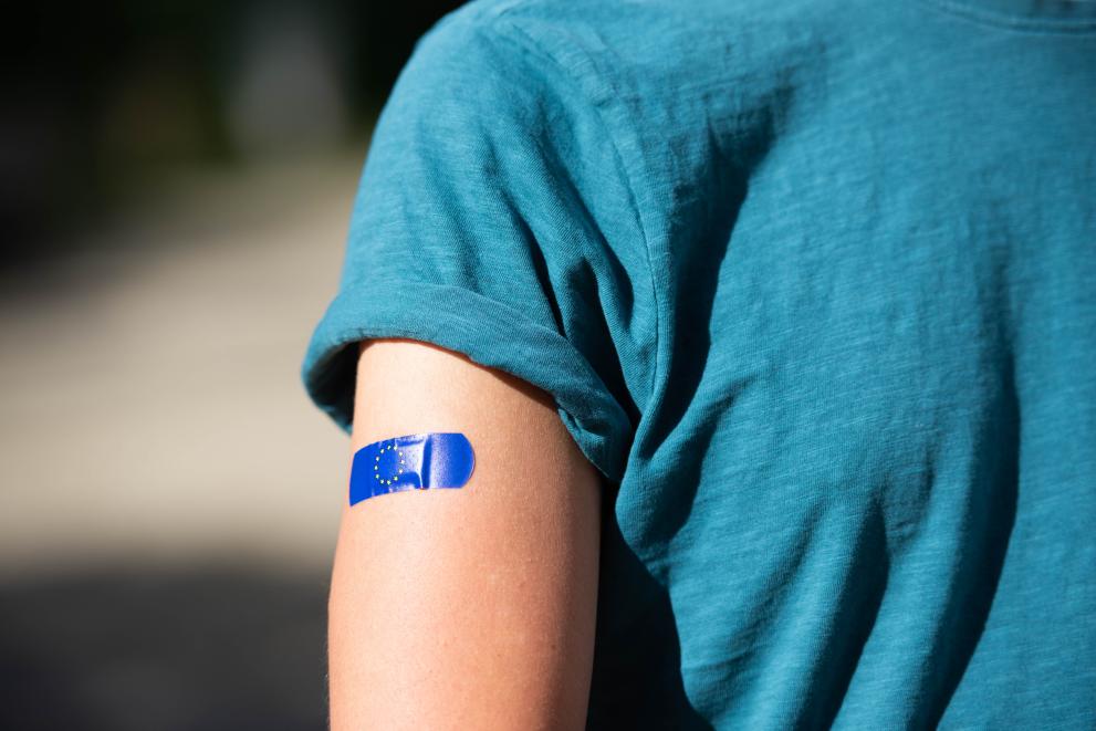 The picture shows half of a womans upper body. Dressed in a blue t-shirt and with a EU-plaster on her arm.