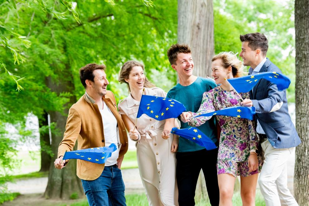 Fünf junge Menschen stehen in einem Park und schwenken kleine EU-Fahnen, während sie lachen.