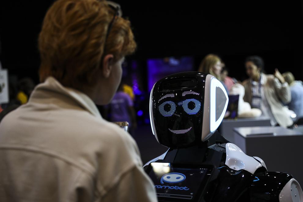 A robot with a digital face (eyes and mouth) is talking to a woman.