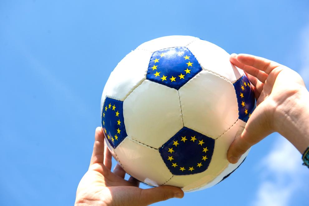   A soccer ball in the colors of Europe is being held up in the sky by two hands.