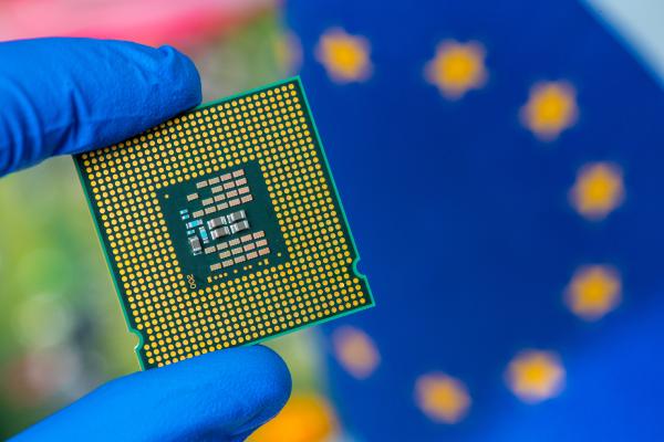 Zwei Finger in blauen Handschuhen halten einen Mikrochip. Im Hintergrund ist eine EU-Flagge zu sehen.